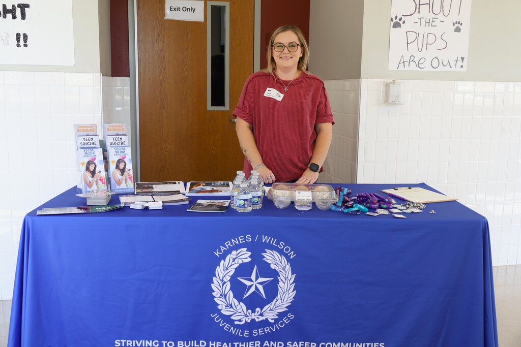 Person in red shirt at a table. Display has books, pamphlets, and items on blue tablecloth. Behind is a door and a sign.