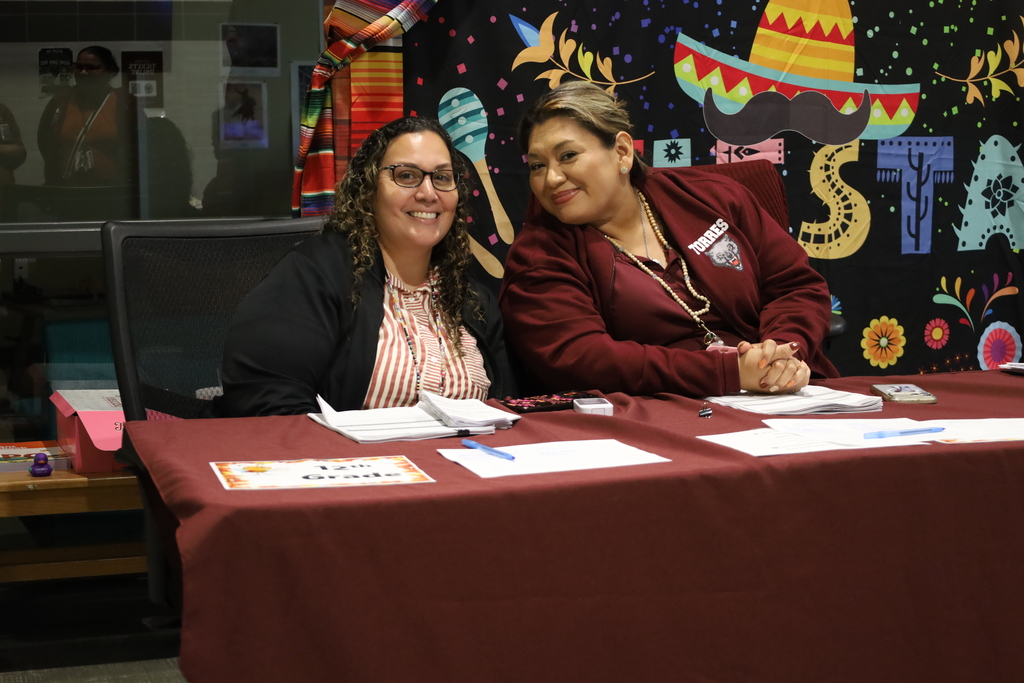 Two women sit at a table with papers and pens, smiling. One wears glasses. A colorful backdrop with 'STA' letters is behind them.