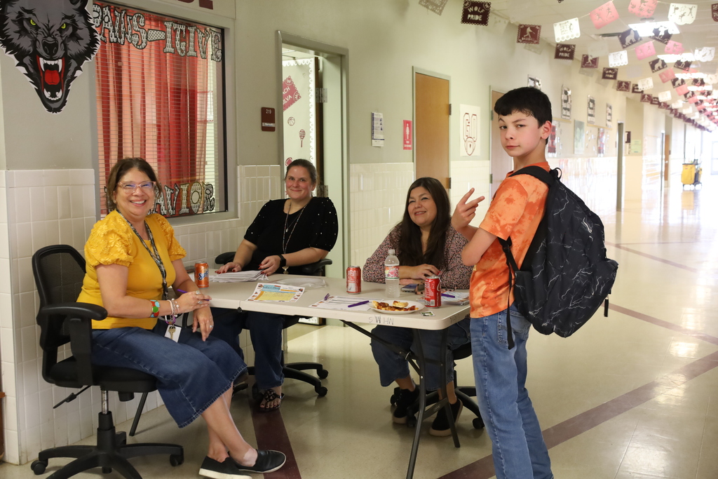 Four people sit at a table in a hallway. A student stands with a backpack. Two women wear glasses.