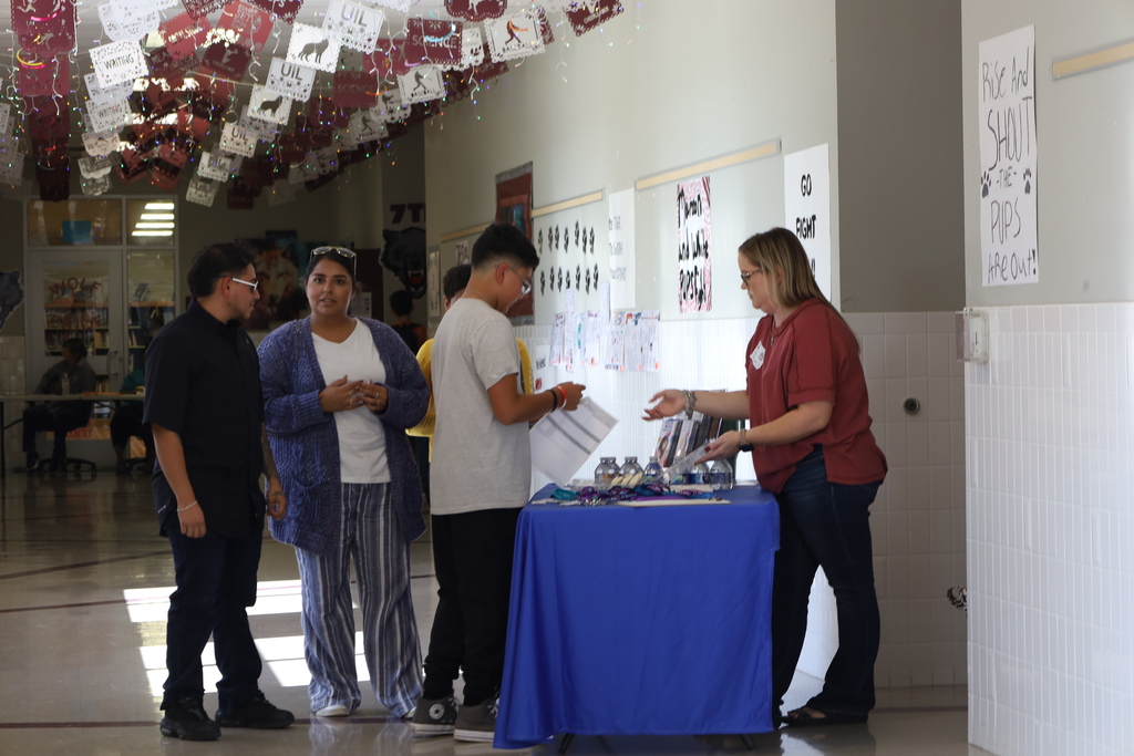 Four people stand around a blue table in a hallway with white walls and bulletin boards.