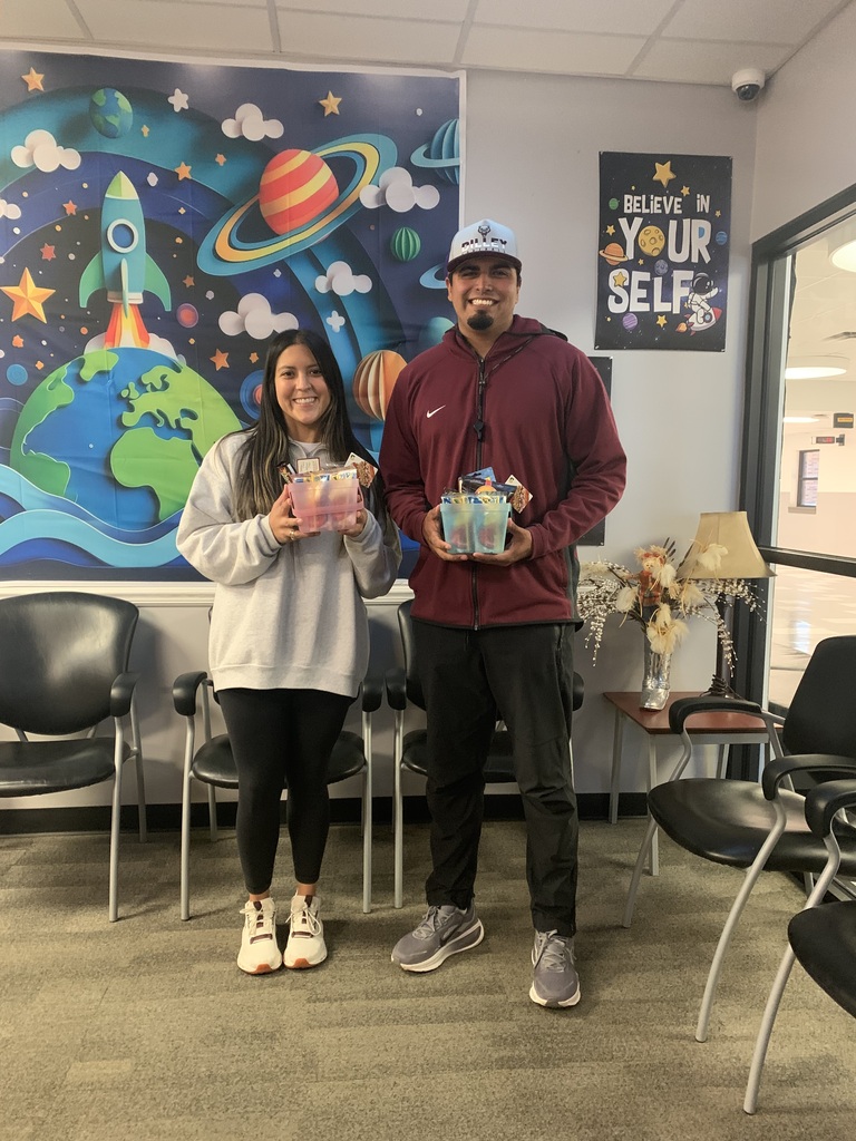 Two people stand next to each other, holding gifts. They are in a room with chairs and a mural.