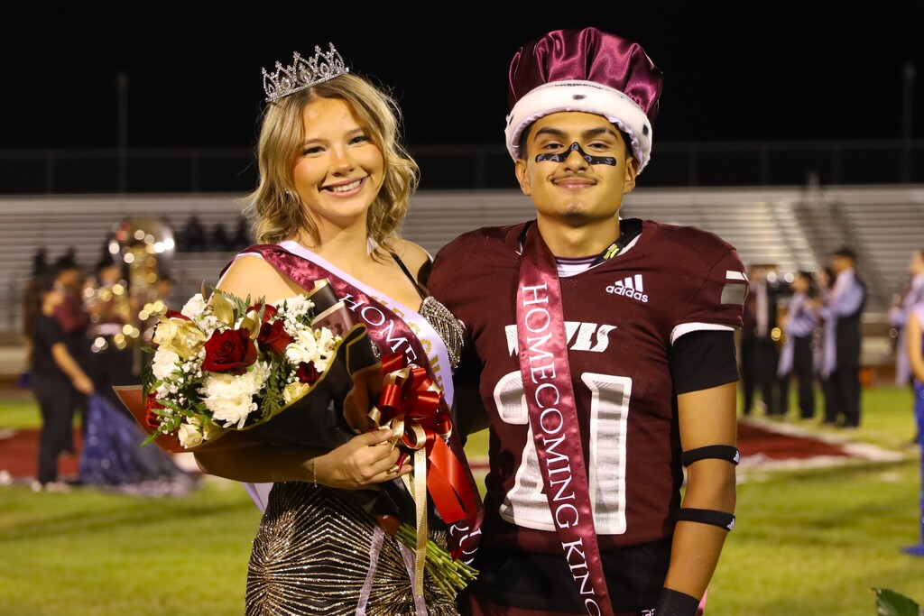 Woman in crown and flowers stands next to a man in maroon jersey, helmet, and sash at a football field.