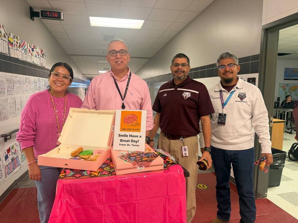 Four people stand around a table in a hallway. One person holds a pizza box. Two men wear white shirts, one wears a pink shirt, and another wears a maroon shirt.