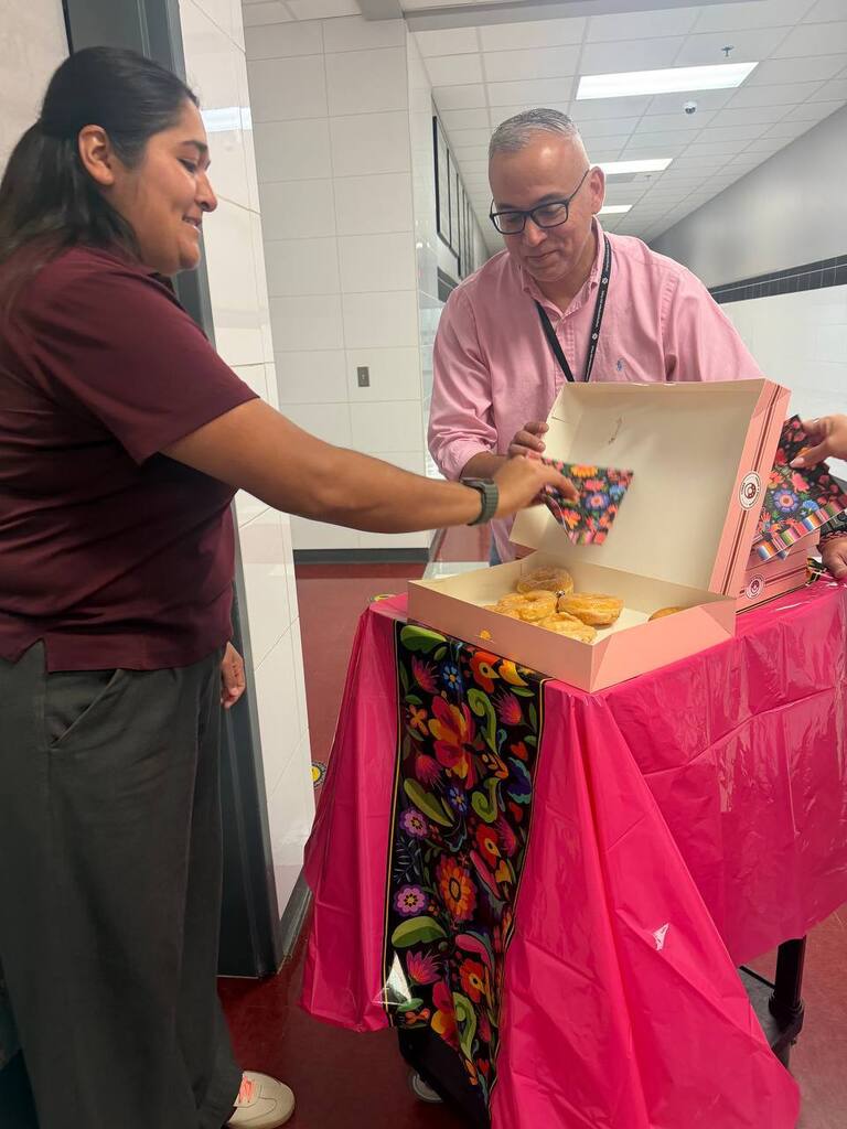 Two adults in a hallway. A person holds a box with pastries. A person hands them a bag.