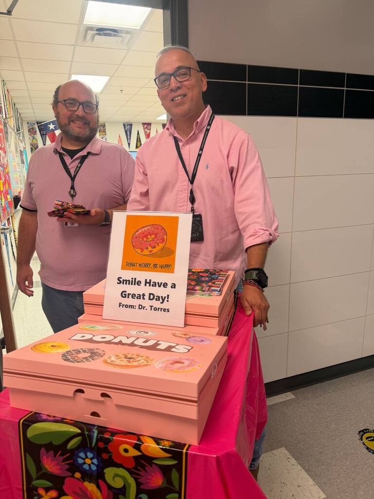 Two men stand behind a table, one holding a sign with a message and a donut box.