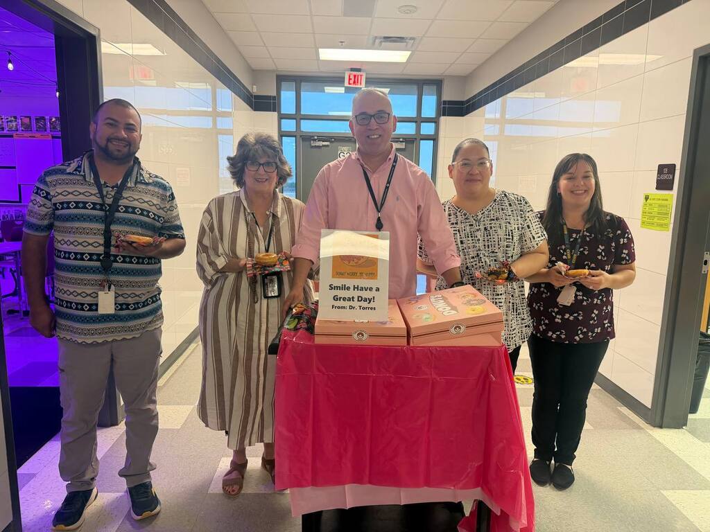 Five people stand in a hallway near a table covered with a pink cloth, holding boxes.