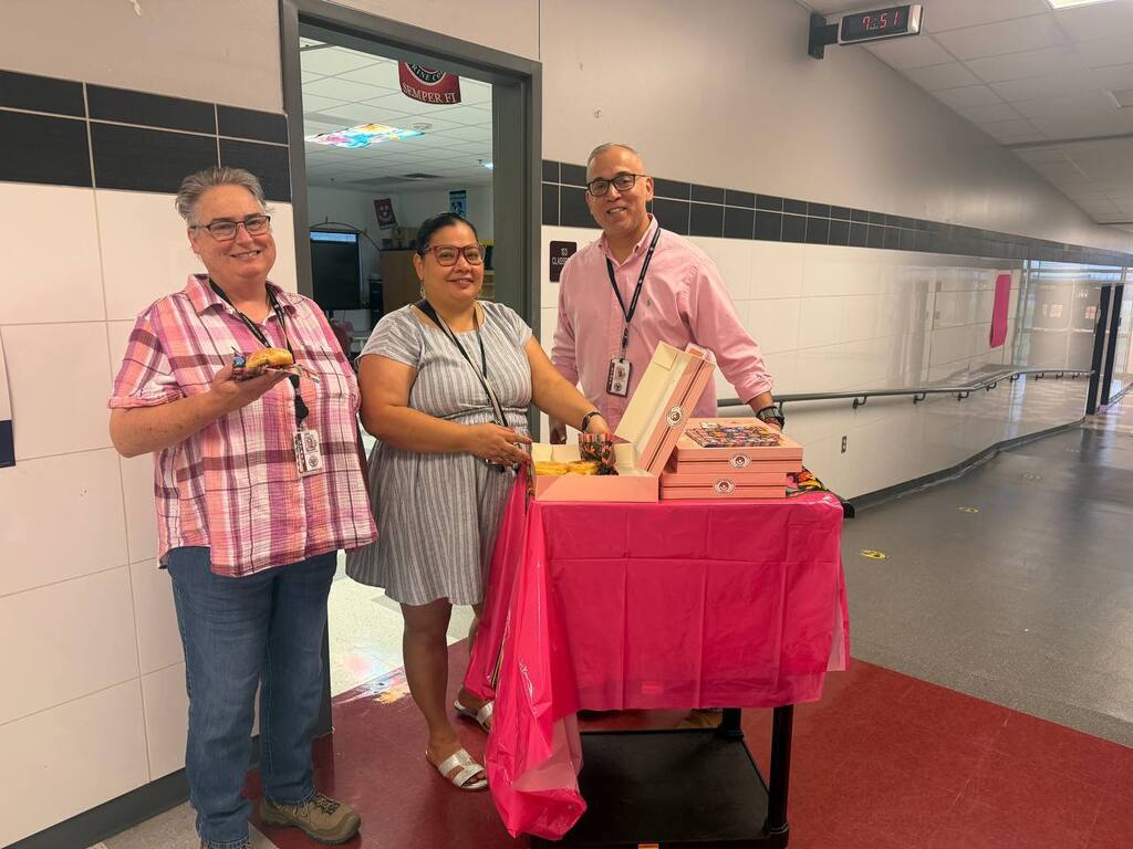 Three adults stand near a pink table, holding food. One person holds a book. They are in a hallway with tiled walls and a red floor.