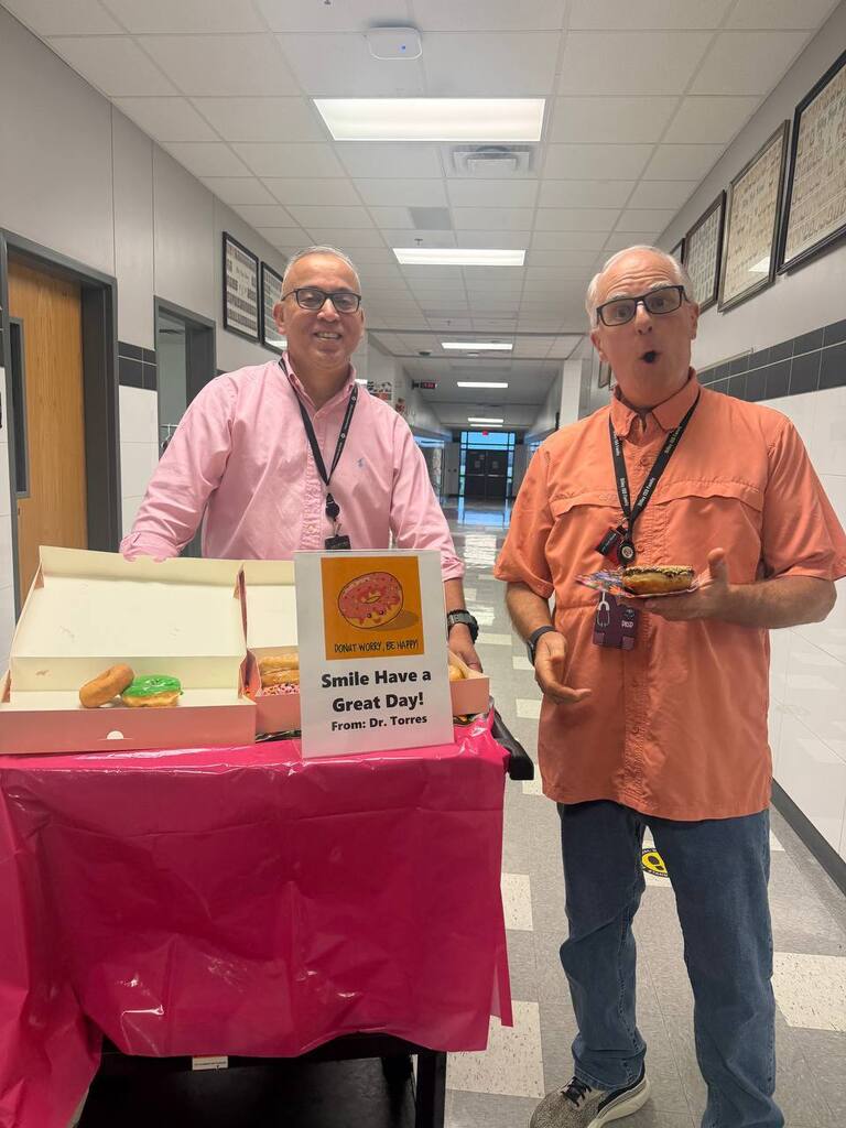 Two men in orange shirts stand by a table with a sign reading "Smile, Have a Great Day!" A pink tablecloth and boxes of food are also visible.