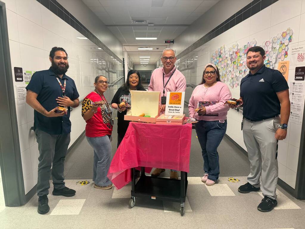 Five people, standing in a hall, pose for a picture. One holds a box, while others hold something. A trolley with a pink cloth is in front of them.