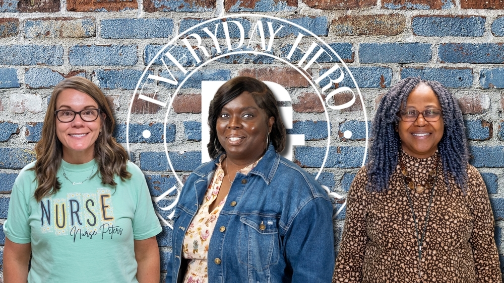 Here is the alt text for the image: Alt text: Three women stand smiling in front of a rustic blue brick wall featuring a circular "Everyday Hero" logo. From left to right: Erin Peters, wearing a mint green "Nurse" t-shirt with her name on it; Chaquilla Green, wearing a denim jacket over a floral top; and Sylvia Frazier, wearing glasses and a brown leopard-print top with blue braided locs.