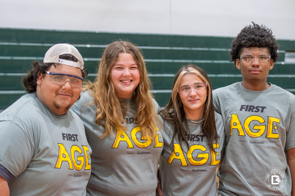 our Woodland High School students wearing gray FIRST Tech Challenge AGI shirts and safety glasses stand side by side in a gymnasium, smiling at the camera with green bleachers in the background.