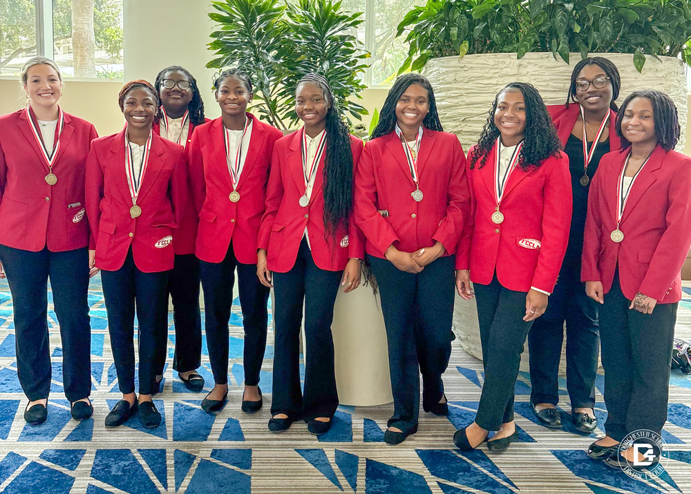 A group of nine Woodland High School FCCLA students stand side by side indoors, smiling and wearing matching red blazers with FCCLA patches, black pants, and medals around their necks. They are positioned in front of large potted plants and a patterned blue carpet.