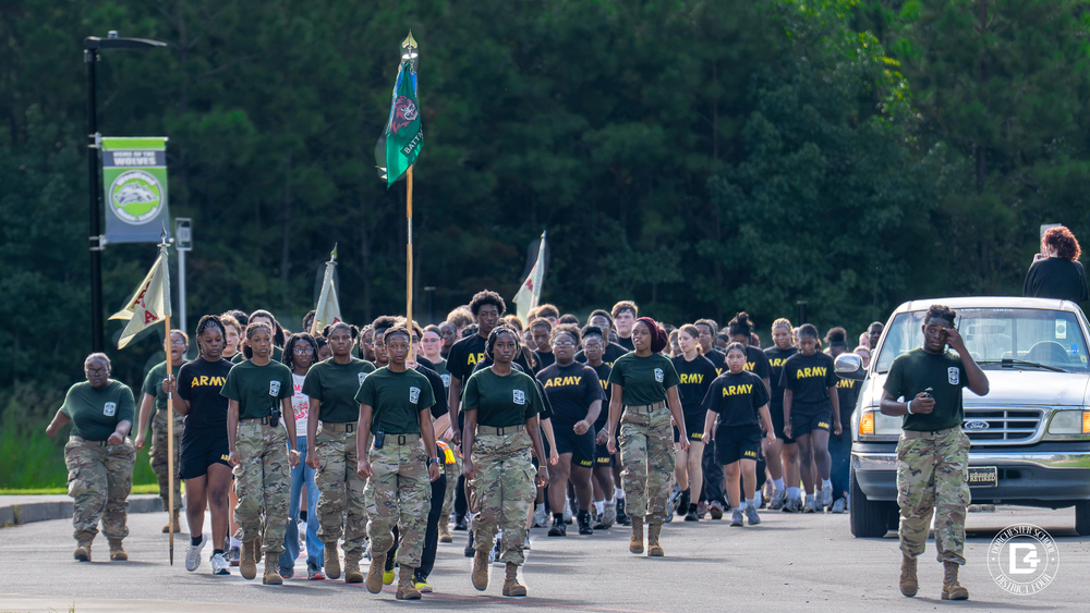 Woodland High School Army JROTC cadets march in formation along a road lined with tall pine trees during Operation Flagpole to Flagpole. Cadets at the front wear green shirts, camouflage pants, and boots, while dozens more behind them wear black Army PT shirts and shorts. One cadet carries a green battalion flag with a red wolverine, and others carry guidon flags. A cadet walks beside a white pickup truck on the right, and a light pole banner reading "Home of the Wolves" marks their arrival at Woodland Middle School.