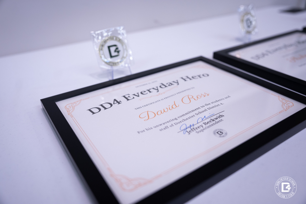 A framed DD4 Everyday Hero certificate on a table recognizes David Ross for his commitment to the students and staff of Dorchester School District Four. A DD4 Superintendent’s Challenge Coin is displayed behind the frame.