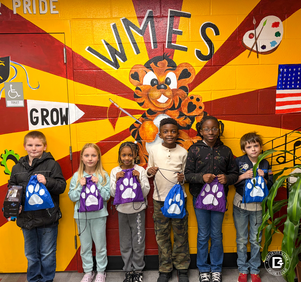 A group of elementary-aged students stand in front of a colorful school mural featuring a tiger mascot and the word “WMES,” each student holding a paw-print drawstring bag.