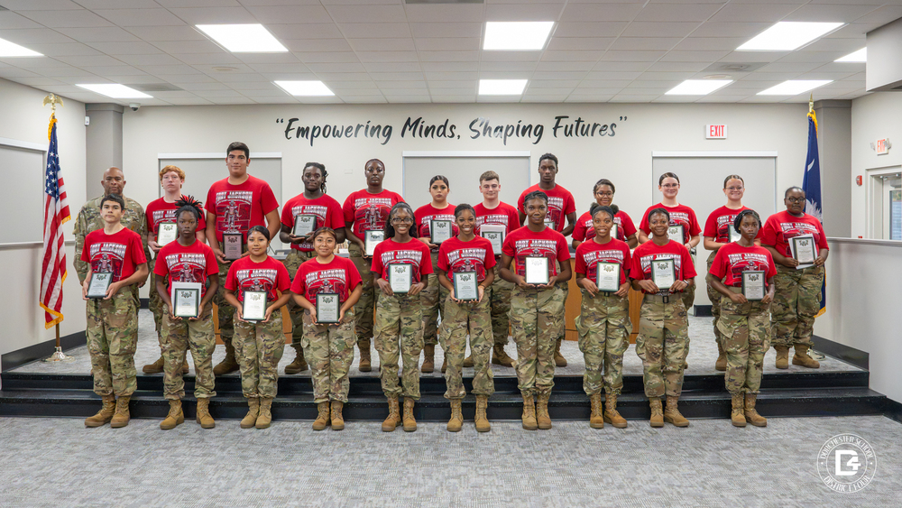 Woodland High School JROTC cadets pose in two rows in the DD4 board room, each holding a plaque. The cadets wear red Fort Jackson JROTC Cadet Leadership Challenge t-shirts with camouflage pants and tan boots. A JROTC instructor in full camouflage uniform stands with them in the back row. The American and South Carolina flags stand on either side, and the wall behind them reads "Empowering Minds, Shaping Futures."