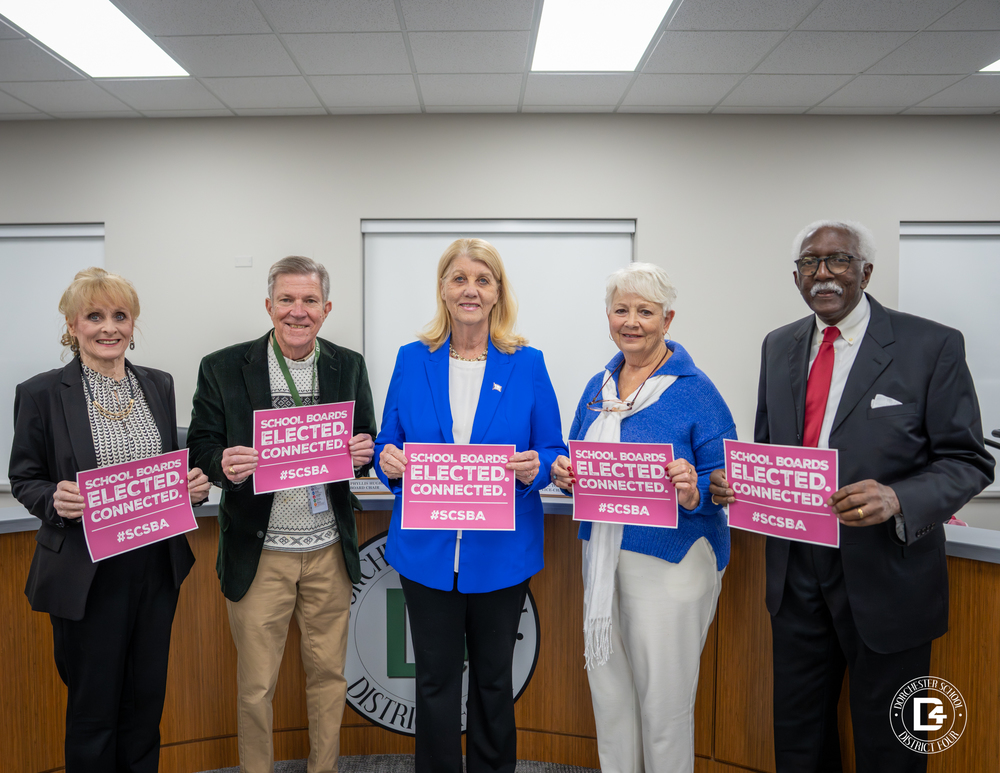 Five Dorchester School District Four Board of Trustees members stand in a meeting room, each holding a pink sign that reads “School Boards Elected. Connected. #SCSBA,” with the district seal visible on the dais behind them.