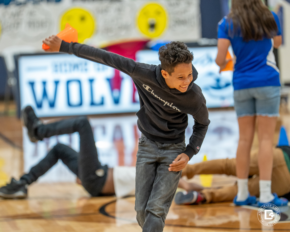 A smiling student in a black hoodie runs forward in the gym while holding an orange cone, with other students playing in the background.