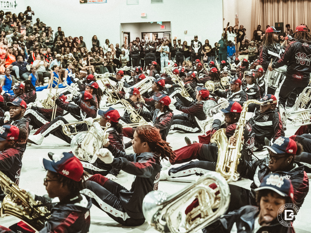 The Marching 100 band, dressed in matching maroon and navy uniforms with red caps, performs seated on the gym floor with brass instruments raised, as a large audience watches.