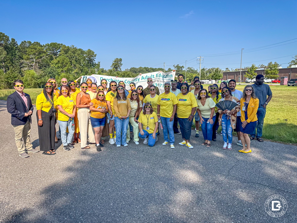 A large group of Dorchester County Adult Education staff and community members stand together outdoors for a group photo. Most are wearing bright yellow shirts and sunglasses, smiling in front of a banner for Dorchester County Adult Education. The background shows trees, grass, and a clear blue sky."