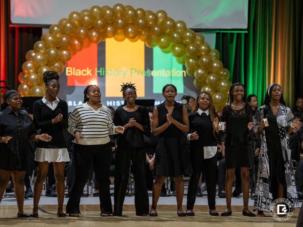 A group of Woodland High School students stand on stage performing during a Black History Month program. The students are dressed in black and sing together beneath a large gold balloon arch. Behind them, a screen displays the words “Black History Presentation” with red, yellow, and green lighting in the background.