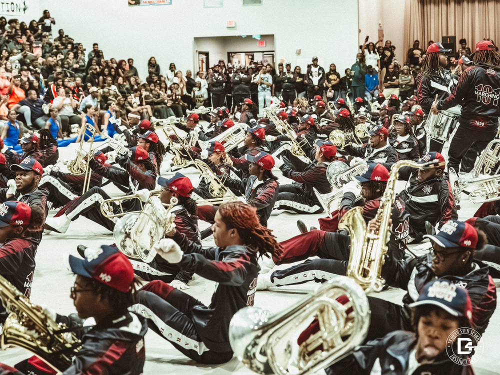 The Marching 100 band, dressed in matching maroon and navy uniforms with red caps, performs seated on the gym floor with brass instruments raised, as a large audience watches.