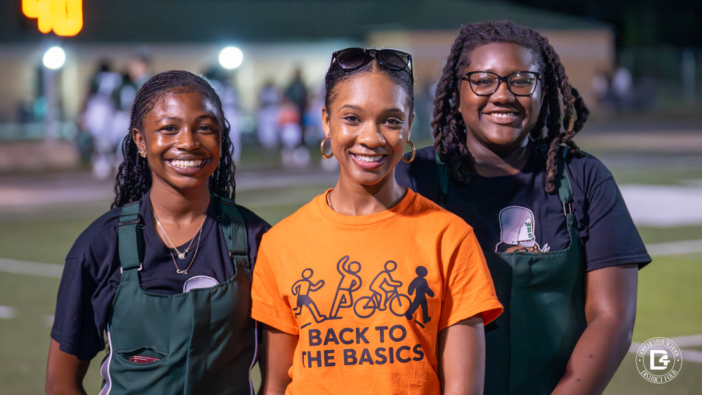 Three young women stand together on a football field at night. The student in the center wears an orange “Back to the Basics” T-shirt with icons of people running, cycling, and walking. She is smiling and wearing hoop earrings and sunglasses on her head. On either side are two Woodland High students in green band uniforms, both smiling brightly.