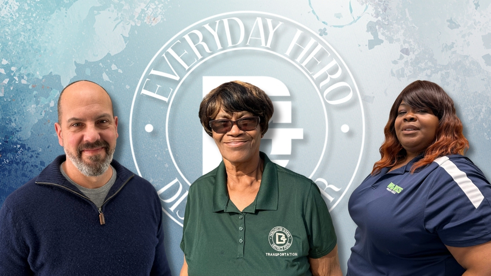 Three adults stand shoulder to shoulder in front of a blue “Everyday Hero” backdrop. Mr. Samson stands on the left wearing a navy sweater, Mrs. Lewis stands in the center wearing a green transportation polo shirt and glasses, and Mrs. Brown stands on the right wearing a navy shirt.
