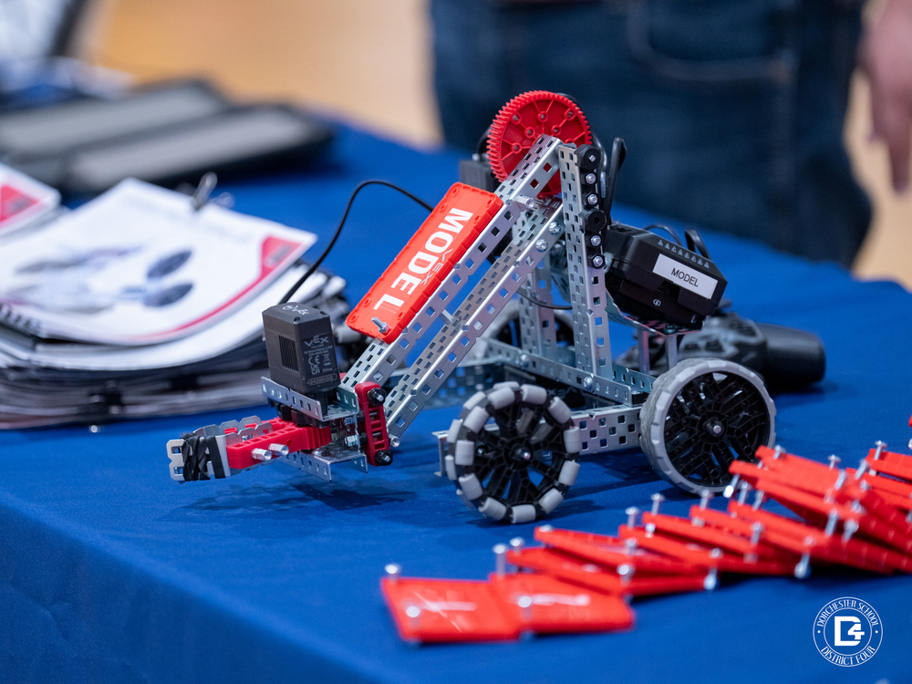 A student-built VEX Claw Bot sits on a blue table during Google’s Robotics Workshop at Woodland Middle School. The robot, labeled “MODEL,” features red and silver metal components, gears, and wheels used for the hands-on STEM activity. Dorchester School District Four’s logo is visible in the corner.