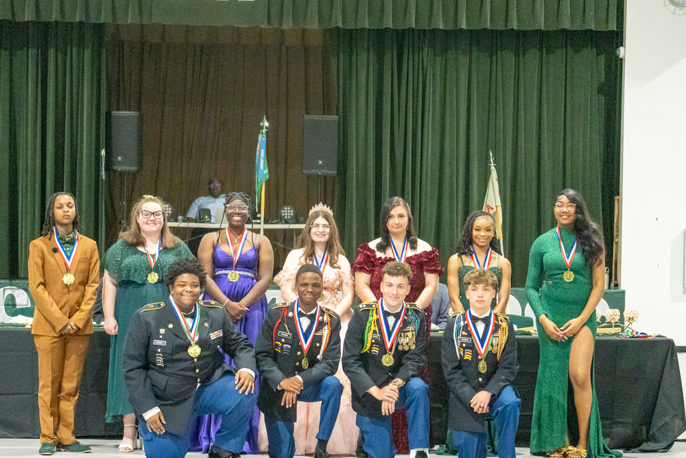 Group photo of Woodland High School JROTC students at the Military Ball. Seven students stand in the back row dressed in formal attire — including evening gowns and dress suits — each wearing gold medals on ribbons. One student in the center wears a tiara. Four students kneel in the front row wearing military dress uniforms adorned with medals, ribbons, and insignia. Two military flags are visible in the background near a stage with green curtains and speakers.