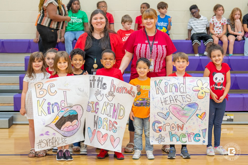 A group of Clay Hill Elementary students stand with two teachers in the gym, smiling and holding colorful handmade posters. The signs read “Be Kind,” “Little Hands Big Kind Words,” and “Kind Hearts Grow Here!” with hearts, flowers, and bright decorations.