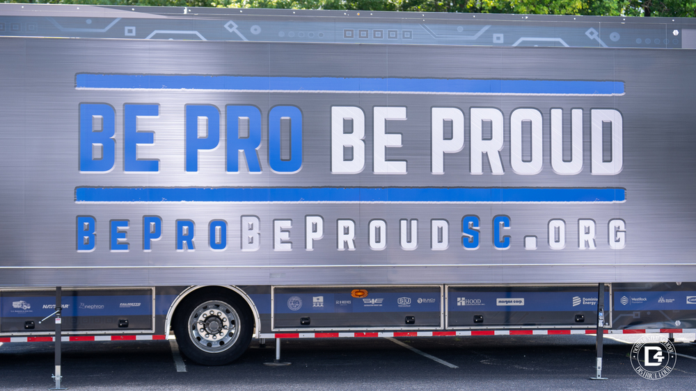A large silver semi-truck trailer with the words “BE PRO BE PROUD” in bold blue and white letters across the side. Below, the website “BeProBeProudSC.org” is displayed. The trailer is part of the Be Pro Be Proud mobile workshop tour, featuring sponsor logos along the bottom panel.