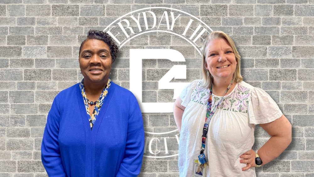 Two women stand side by side in front of a gray brick wall featuring a large Dorchester School District Four “D4” logo and the words “Everyday Hero” in a circular design. The woman on the left wears a royal blue sweater and a patterned blouse, smiling softly at the camera. The woman on the right wears a light-colored blouse with floral embroidery, a lanyard with keys and ID badge, and smiles with one hand on her hip.