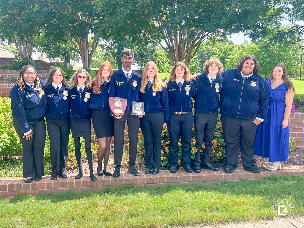 Woodland High FFA students stand outdoors in official FFA jackets, proudly holding awards. Their advisor, Markie Leonard, stands to the right wearing a blue dress. The group is smiling in front of a landscaped brick wall and flowering plants.
