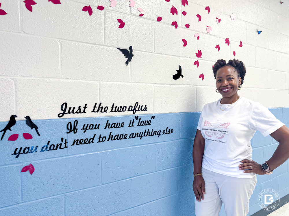 TJ Cheeks, owner of Cheeks Daycare Academy, stands smiling beside a decorated hallway wall at her daycare. The wall features pink and black paper birds and petals with a quote that reads, “Just the two of us. If you have it ‘love,’ you don’t need to have anything else.” She wears a white Cheeks Daycare Academy T-shirt and light-colored pants, standing confidently with one hand on her hip. The Dorchester School District Four logo appears in the lower right corner of the image.