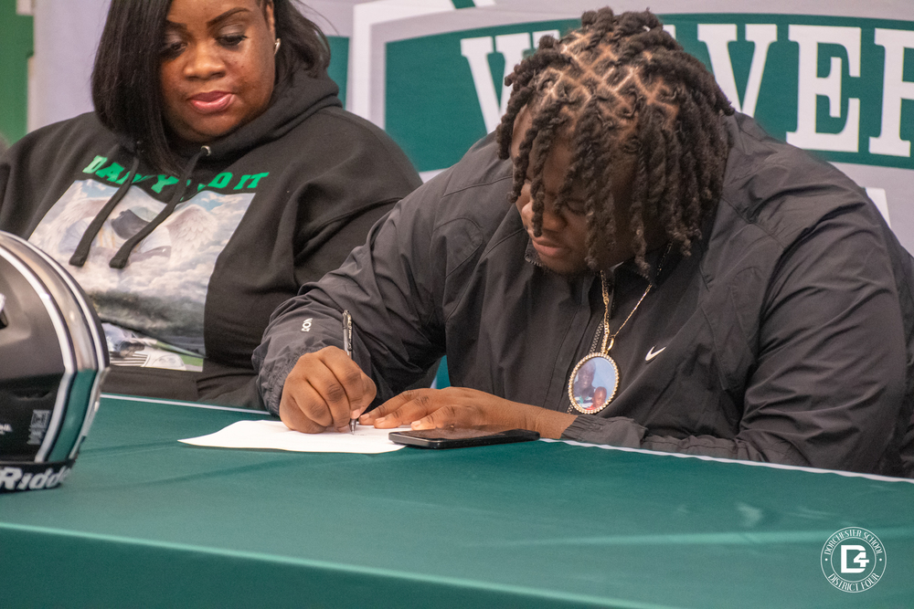 A student athlete sits at a green table signing a document during a college signing ceremony. A Woodland Wolverines football helmet is displayed on the table. Two adults sit beside the student, watching proudly, as a large Wolverines backdrop fills the background. The Dorchester School District Four logo appears in the bottom corner.