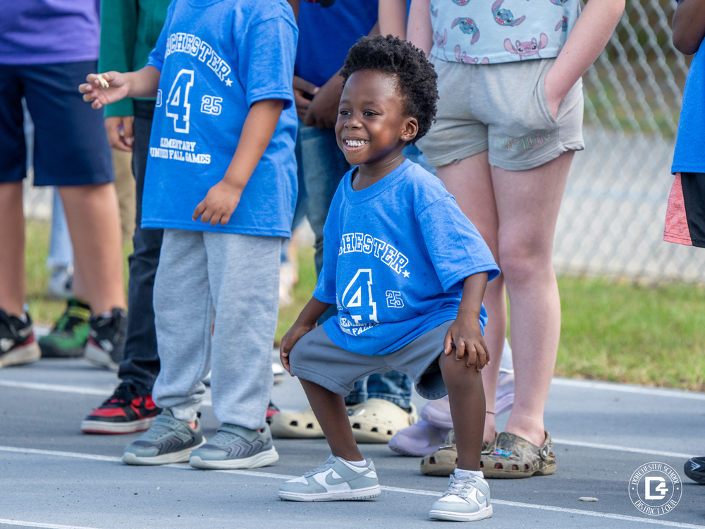 A small child in a blue Dorchester District Four Fall Games shirt crouches playfully with a wide smile while standing with other children near a track.