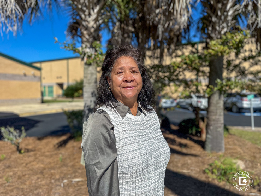 Ms. Shirley Eskew stands outdoors in front of a school building on a sunny day. She is smiling and wearing a gray blouse with a light gray patterned vest. Behind her are palm trees, greenery, and parked cars, with the Dorchester School District Four logo visible in the bottom right corner of the image.
