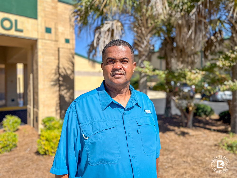 David Ross stands outside a school building on a sunny day, wearing a bright blue button-up shirt. The background features brick walls, green trim, palm trees, and landscaped shrubs, with clear blue skies overhead. The Dorchester School District Four logo appears in the bottom right corner of the image.