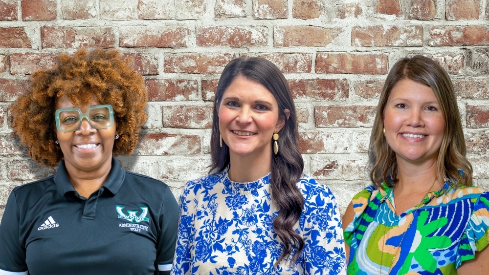 The image shows three women standing in front of a light red and white brick wall. On the left is Ms. Tamara Rolack wearing a black Woodland Administrative Staff polo and large square green glasses, smiling with natural curly hair. In the center is Mrs. Carlie Nelson wearing a white blouse with a bright blue floral pattern, long dark hair, and gold earrings. On the right is Mrs. Angela Crosby wearing a colorful blue, green, and orange patterned blouse, smiling with shoulder length light brown hair.