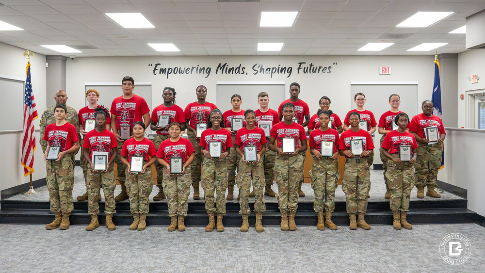 Woodland High School JROTC cadets pose in two rows in the DD4 board room, each holding a plaque. The cadets wear red Fort Jackson JROTC Cadet Leadership Challenge t-shirts with camouflage pants and tan boots. A JROTC instructor in full camouflage uniform stands with them in the back row. The American and South Carolina flags stand on either side, and the wall behind them reads "Empowering Minds, Shaping Futures."