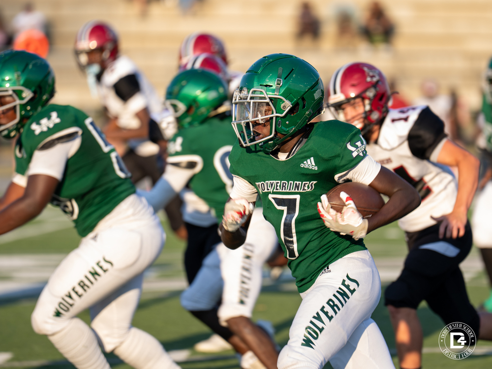 Woodland JV Wolverines football player number 7 in a green uniform runs the ball upfield during the home opener against Bamberg-Ehrhardt, with teammates blocking and Raiders defenders in pursuit. The action takes place under bright evening sunlight on the football field.”