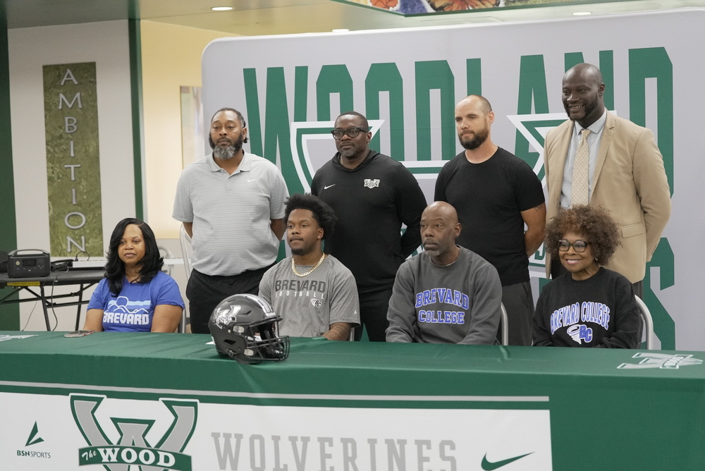 Isaiah Barnes sits at a green-draped signing table alongside family members, all wearing Brevard College apparel, during his college commitment ceremony at Woodland High School. A Woodland Wolverines football helmet rests on the table in front of them. Four coaches and school staff stand behind the group, with a large Woodland Wolverines banner displayed in the background.
