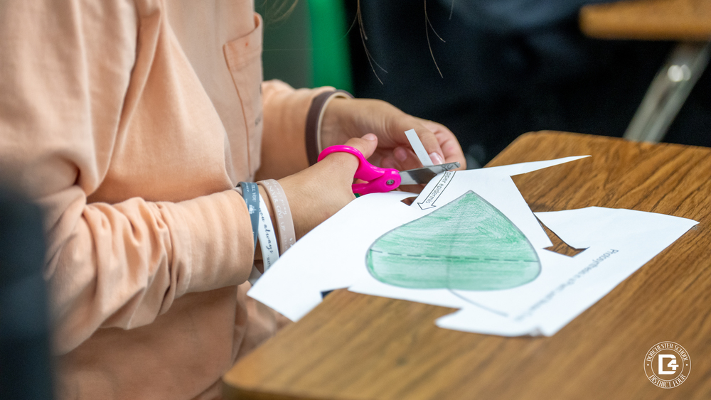 Close-up of a student with pink scissors cutting out a large, green-colored paper leaf for the classroom model.
