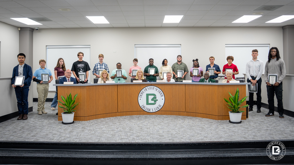 Dorchester School District Four Board members sit at the front table while a group of Woodland Middle and High School students stand behind them holding framed certificates. The students were recognized for outstanding academic achievement at the September board meeting.”