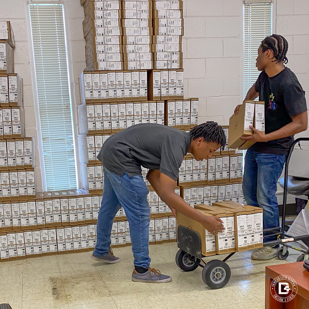 Two young men from the DD4 Summer Work Program load Dell Chromebook boxes onto a dolly in a room filled with stacked boxes. Sunlight filters through vertical blinds behind them.