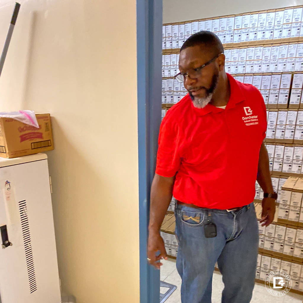 Mr. DeLee from the DD4 Technology Department, wearing a red district polo shirt, walks into a room packed wall-to-wall with Dell Chromebook boxes.