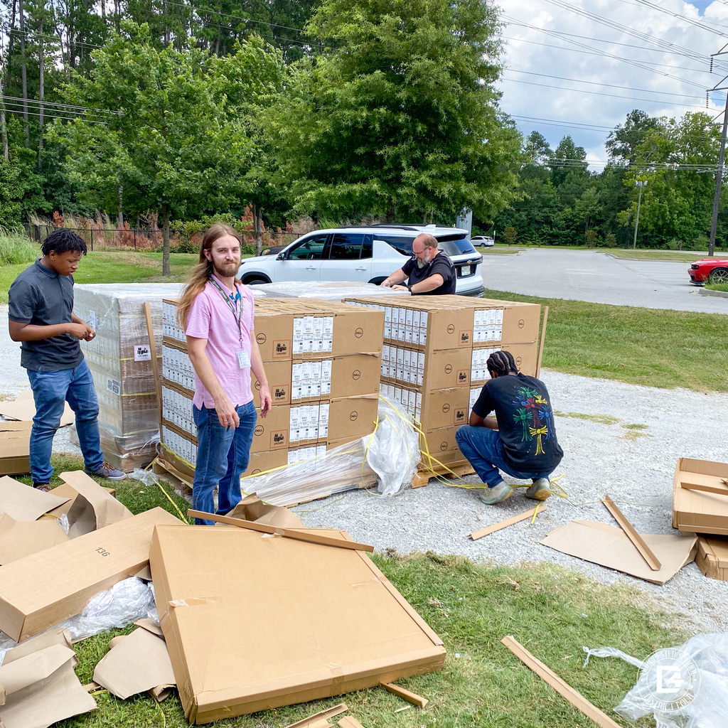 Members of the DD4 Technology Department and Summer Work Program unload Chromebook shipments outside, surrounded by pallets and cardboard in a parking area near trees.