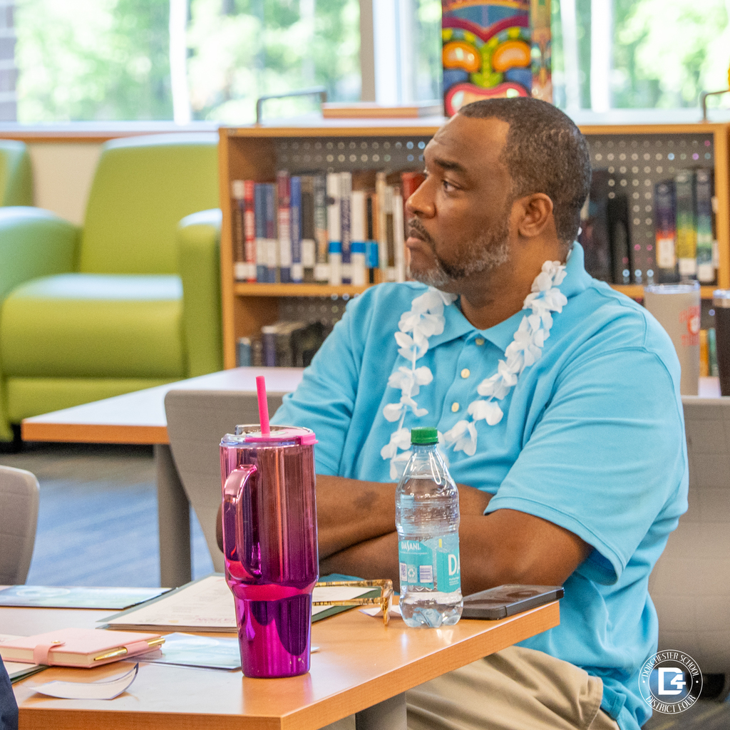 A man wearing a blue shirt and white flower lei listens intently during orientation activities in the school media center.