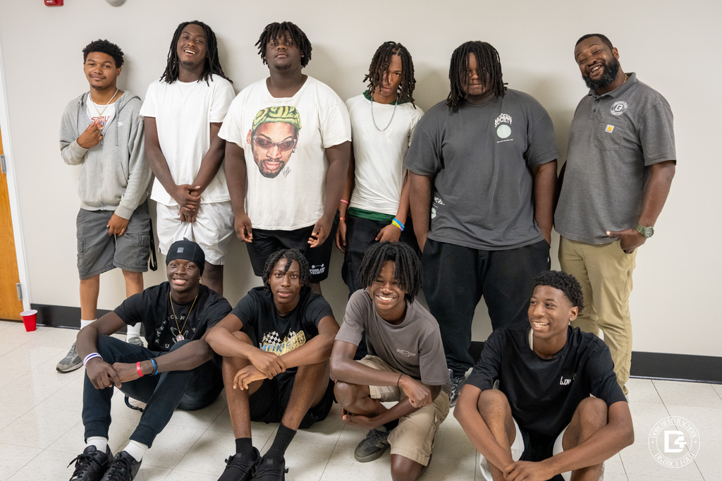 A group of young men and their supervisor from the Dorchester School District Four Summer Work Crew pose together and smile for a group photo inside a school hallway. Some are standing while others are seated in front of them.